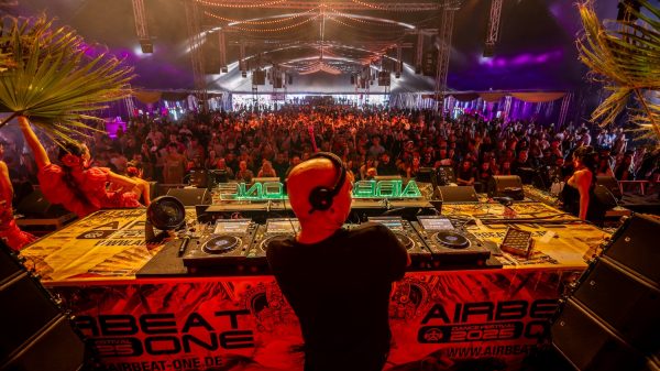 DJ STBAN performing an Afro-Tribal House set on stage at a festival, with dancers and the AIRBEAT ONE logo visible on the DJ booth.